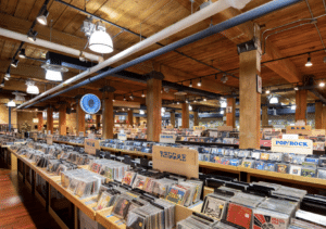 The interior of a large music store, with wooden beams and ceiling, invites you to explore your story among rows of CDs and albums. The genres are neatly labeled—Reggae, Pop/Rock, and more—waiting to become the soundtrack of your next chapter.